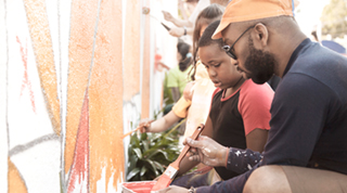Un père noir et son fils participent à la peinture d’une murale colorée sur le pan d’un immeuble.