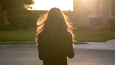 Une femme vue de dos admire le soleil qui se couche derrière une maison située sur une rue de banlieue.